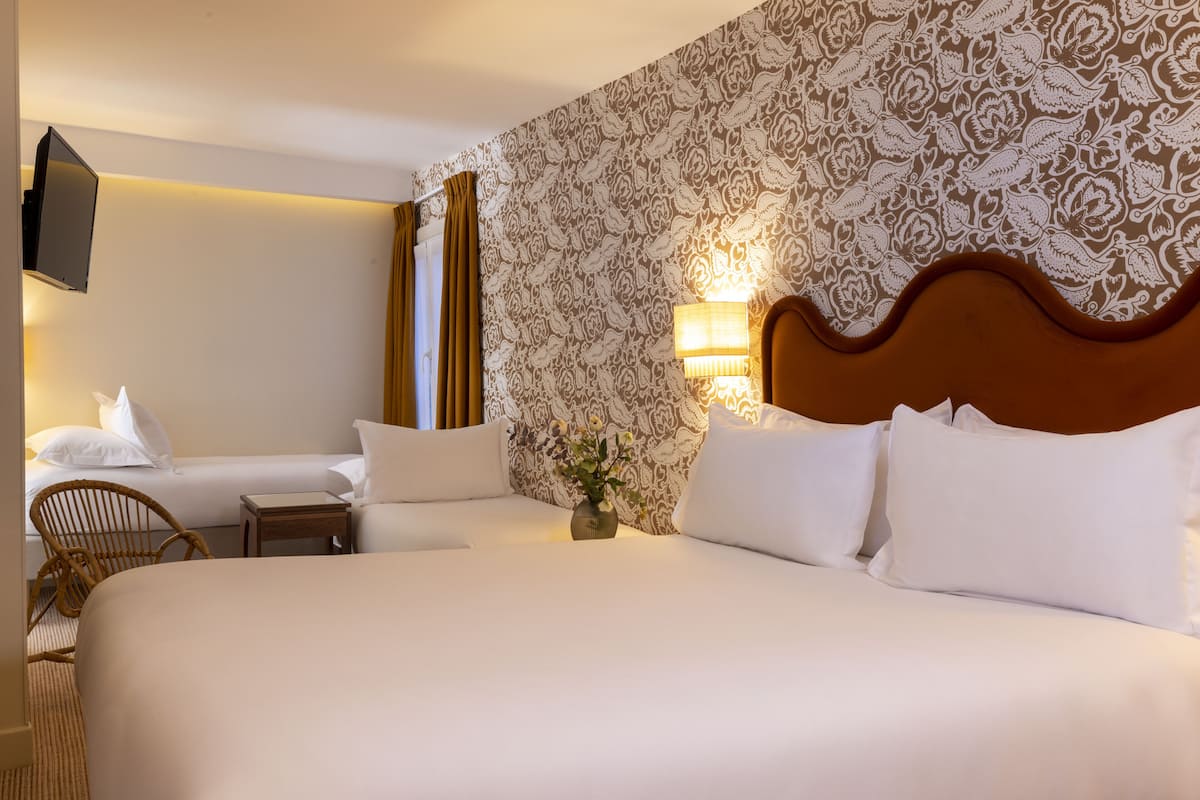 A hotel room with a large bed, a smaller bed, a television mounted on the wall, a lamp, a chair, and a patterned wallpaper.