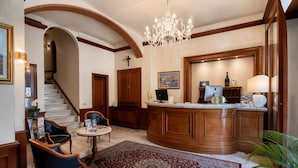 A lobby with a reception desk, a chandelier, a staircase, and a patterned rug. - Hotel Posta (Cecina)