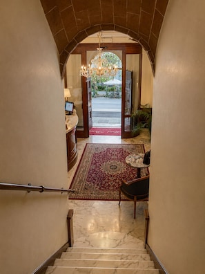 Una escalera de mármol conduce a un vestíbulo con candelabro, alfombra roja y vistas al exterior.