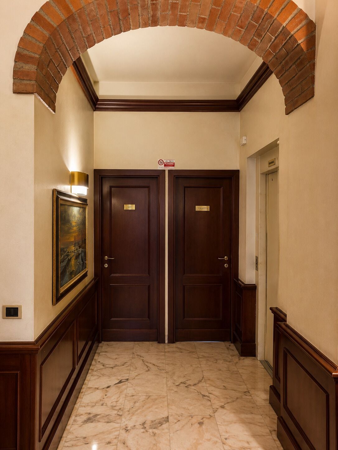 Een gang met twee deuren, aangeduid als '101' en '102', die leiden naar een badkamer, een lijst met een schilderij aan de linker muur, en een bakstenen boog bovenin.