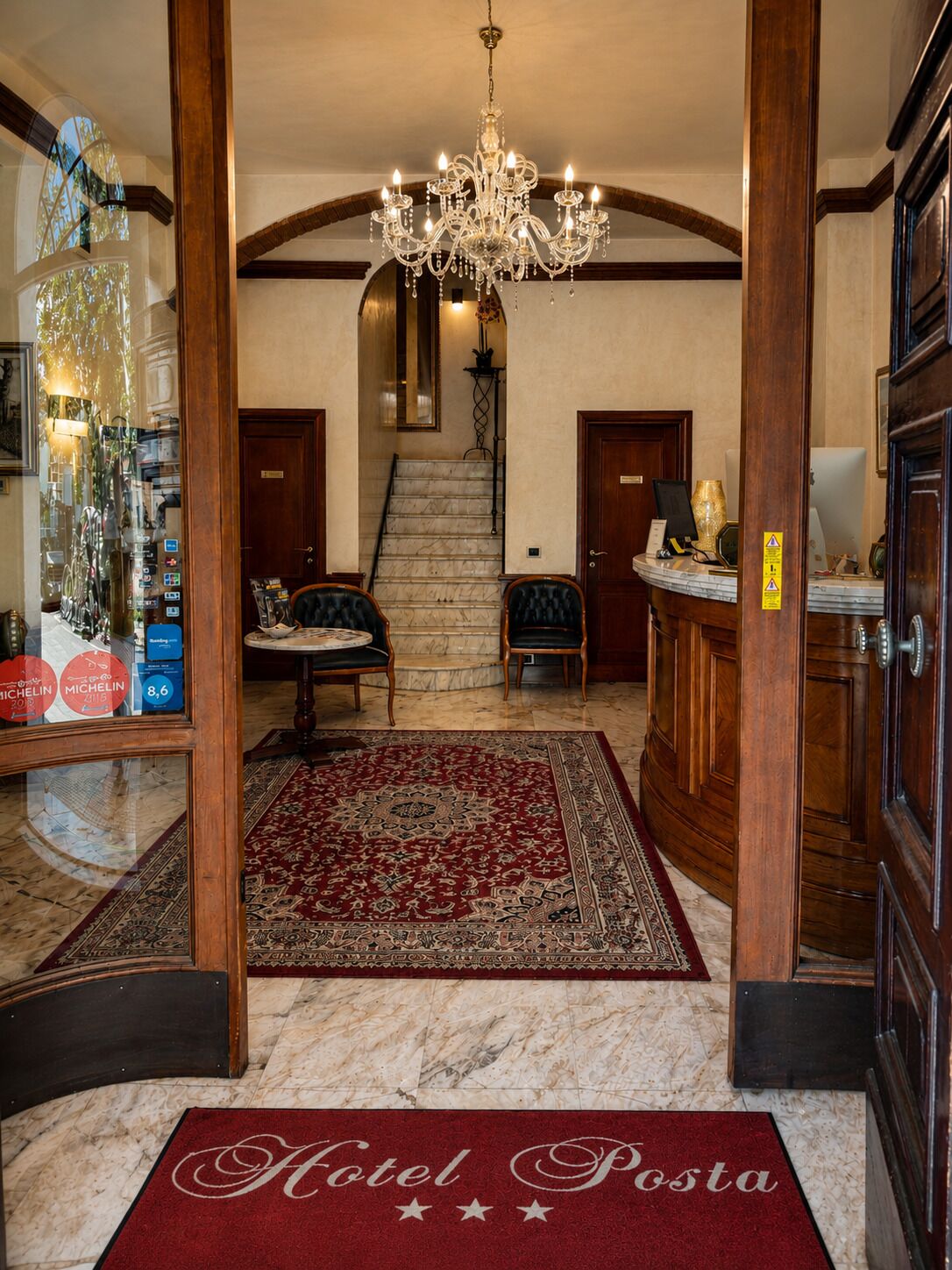 Un'hall d'hotel con un lampadario, una scalinata, una reception e un tappeto rosso con la scritta 'Hotel Posta'.