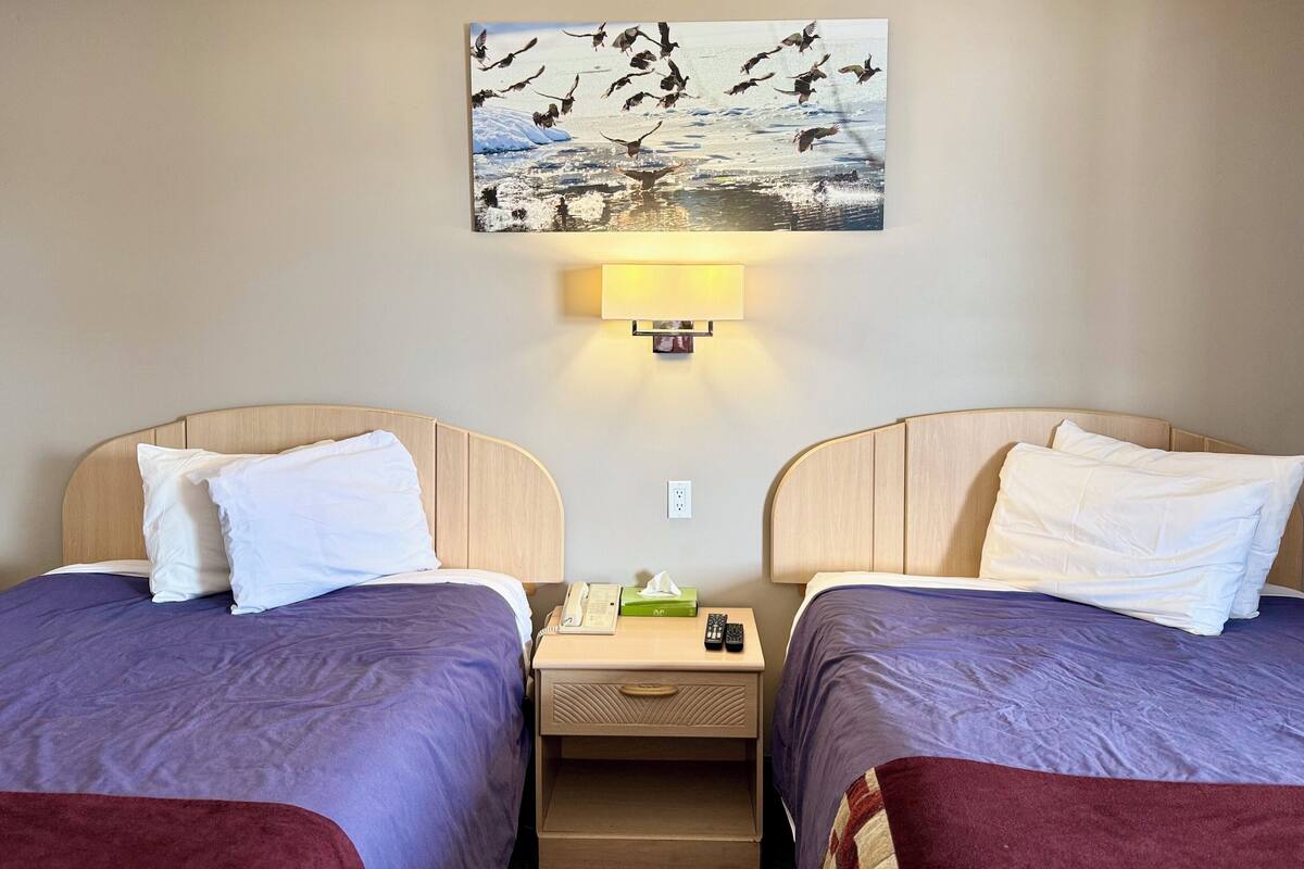 A hotel room with two beds, a nightstand, a lamp, a painting, and a telephone.