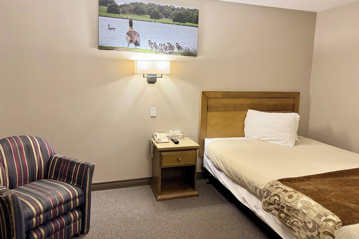 A hotel room with a bed, a chair, a bedside table with a lamp, and a wall-mounted picture of a lake scene.