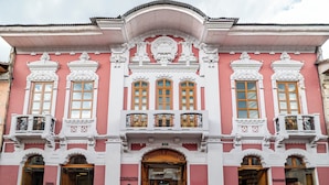 A pink and white building with ornate architectural details, including a central entrance with a wooden door and multiple windows with decorative ironwork. - Carlota Sustainable Design Hotel (Quito)