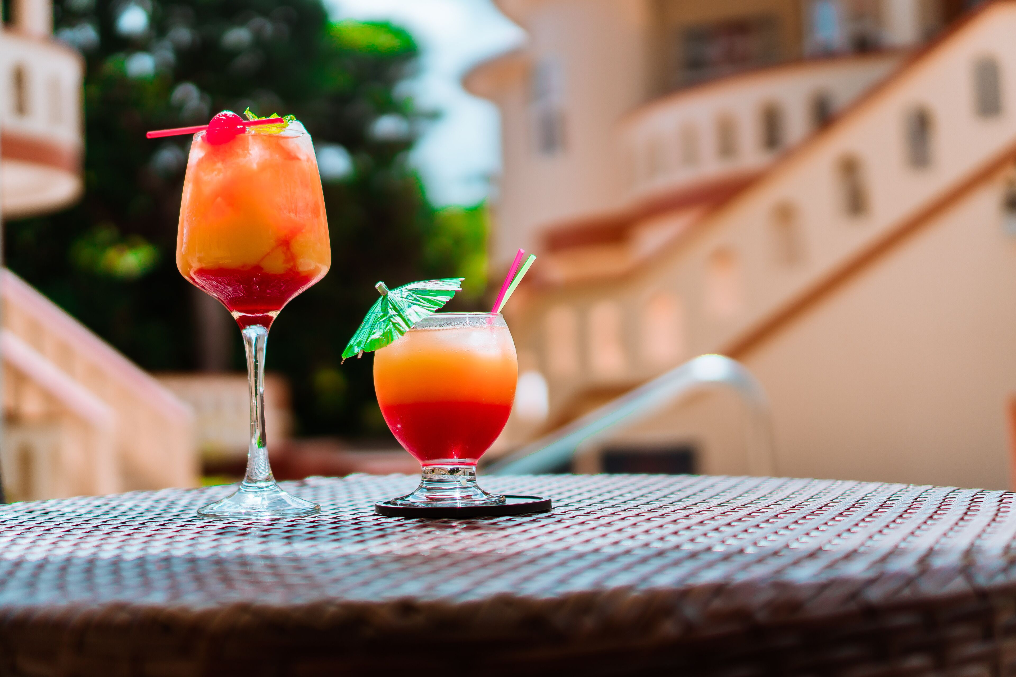 Two cocktails on a table with a woven texture, set against a building with a staircase and greenery in the background.