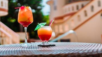 Two cocktails on a table with a woven texture, set against a building with a staircase and greenery in the background.