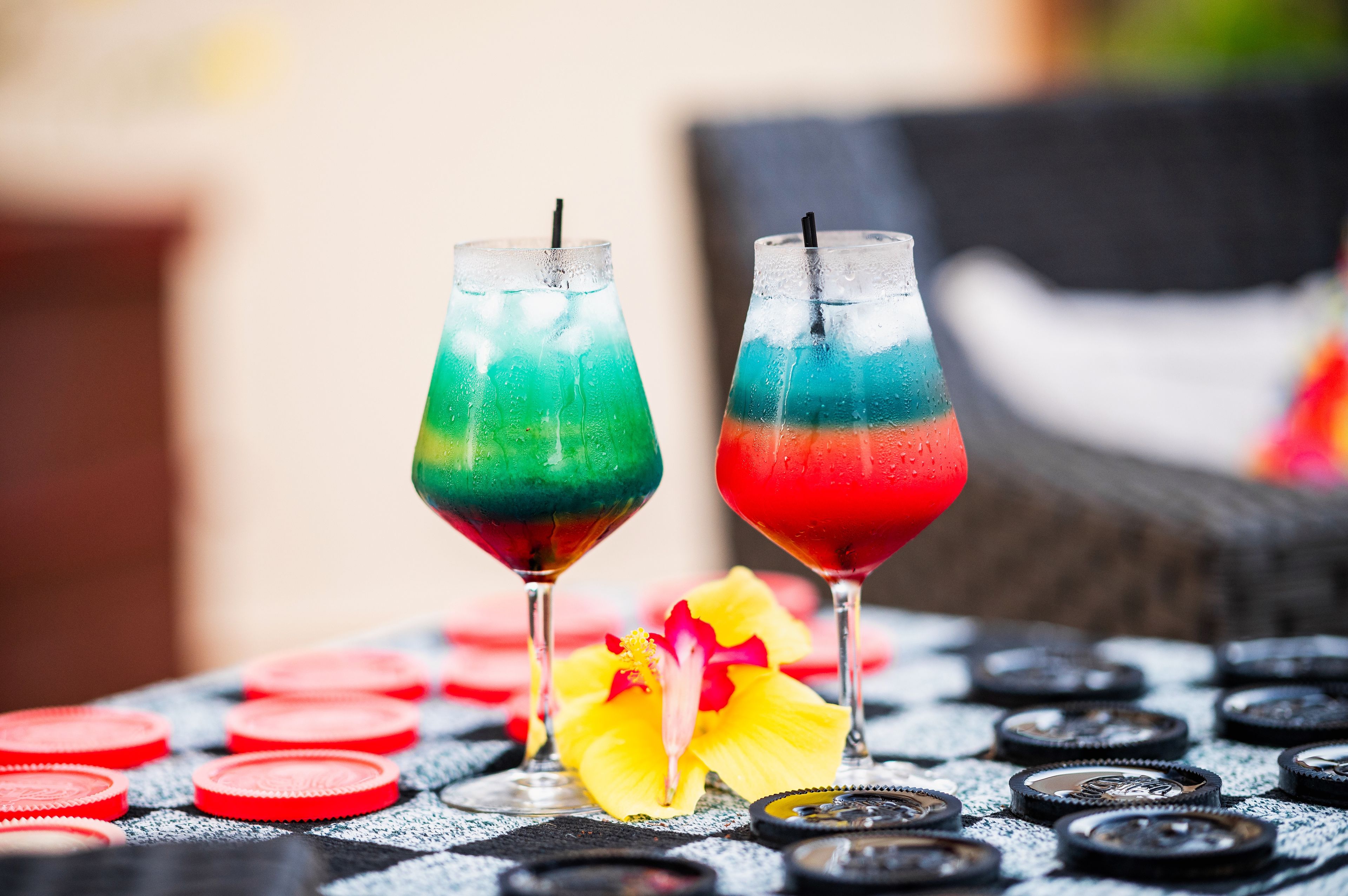 Deux boissons colorées servies dans des verres, posées sur une table avec des jetons de poker et une fleur.