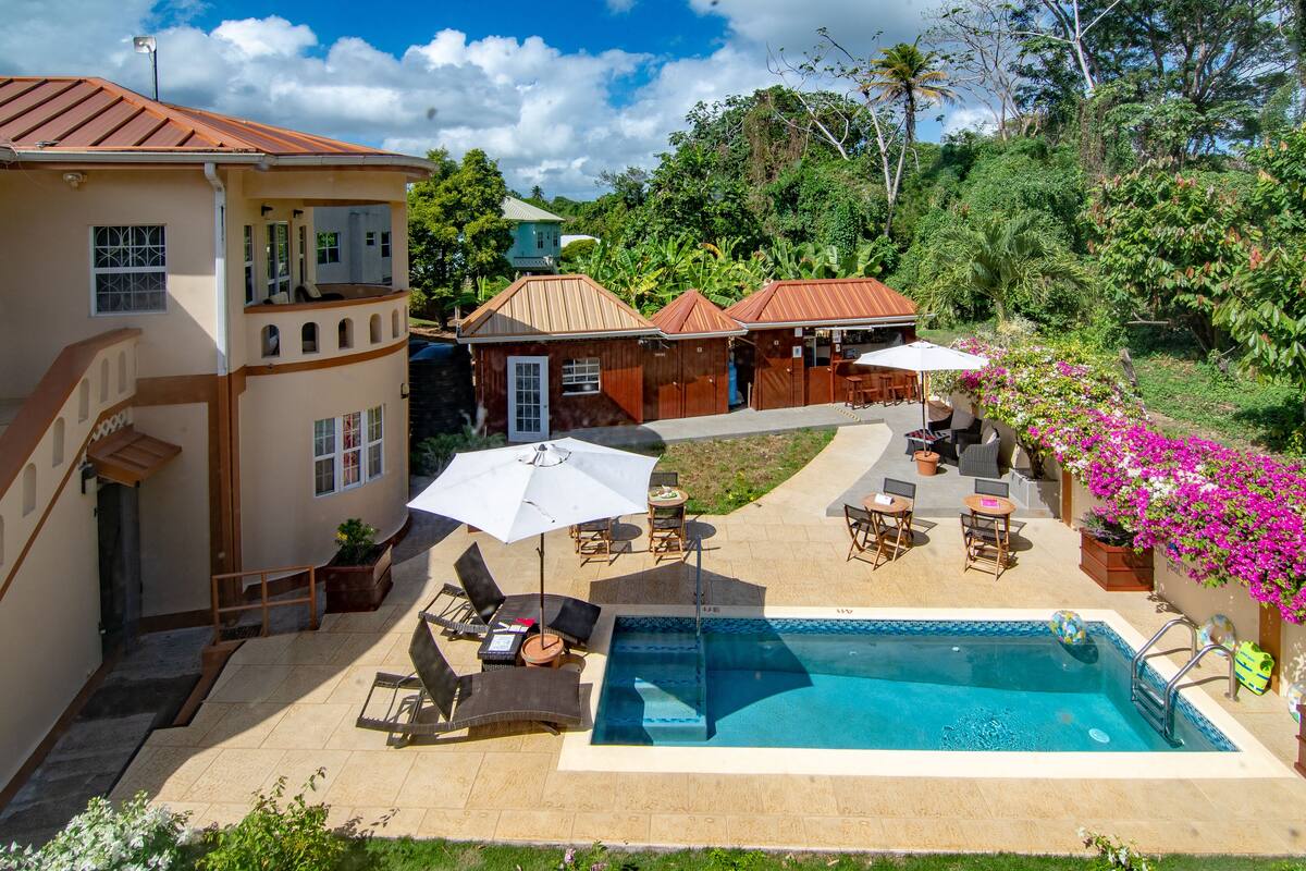A pool area with lounge chairs and umbrellas, surrounded by a house with a red roof and a garden with pink flowers.