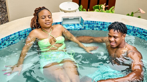 Un couple se détend dans un petit bain à remous carrelé, doté d’un panneau en bois et d’une petite table à proximité.