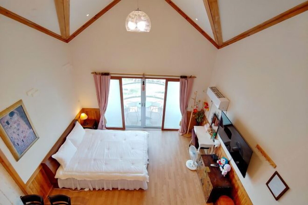 Un dormitorio con una cama grande, un escritorio con una computadora, un televisor y una ventana con cortinas.
