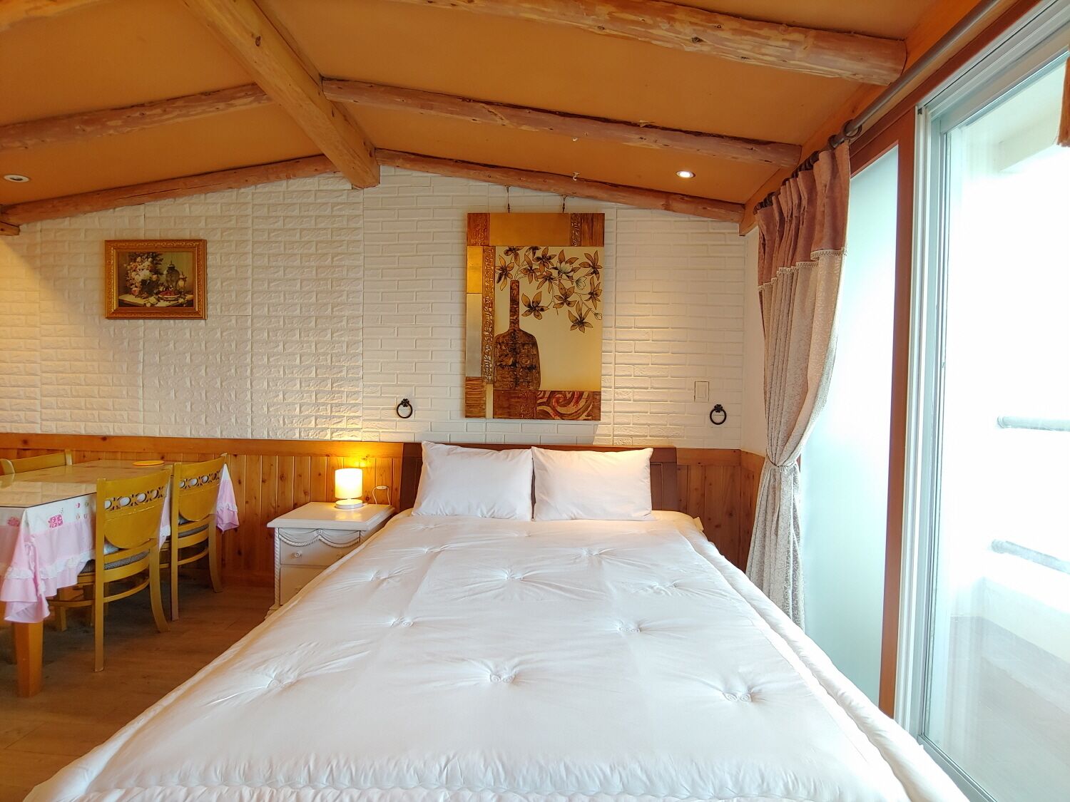 Une chambre avec un grand lit, une tête de lit en bois, un mur en briques blanches, un tableau, une table avec des chaises et une fenêtre avec des rideaux.