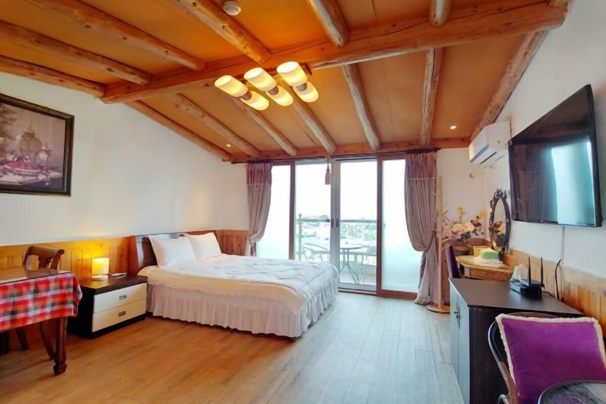 Un dormitorio con techo de madera, una cama, un escritorio con silla, un televisor y un balcón con vistas.