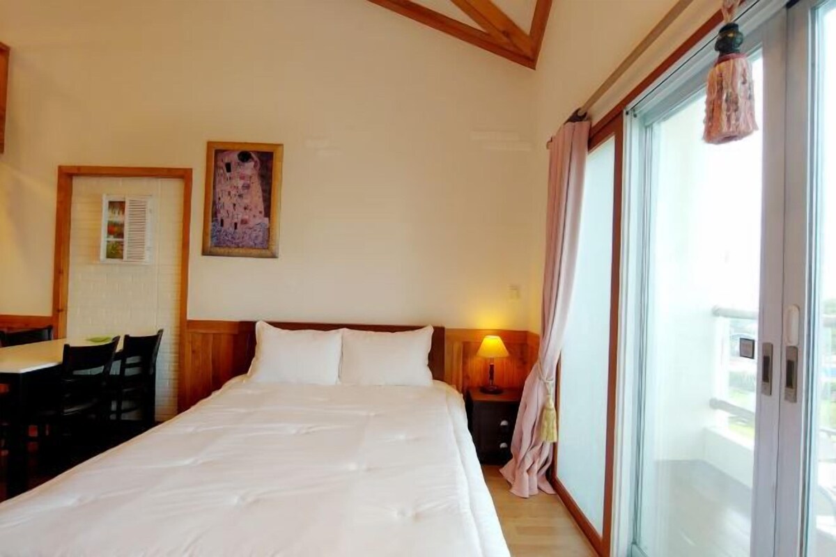 A hotel room with a large bed, a desk with chairs, a painting on the wall, and a balcony with curtains.