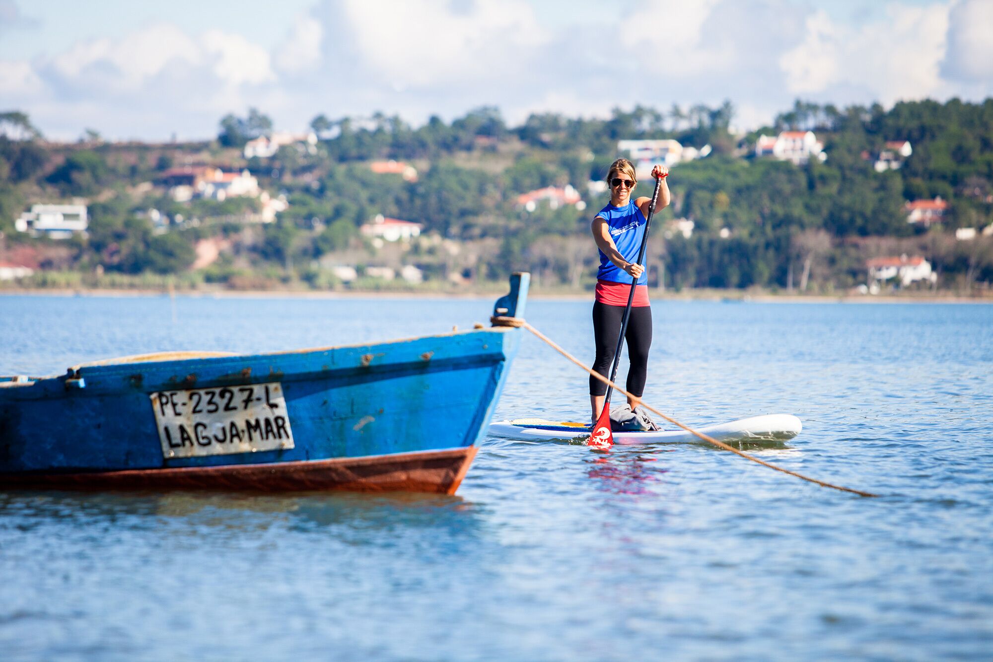 Una persona de pie en una tabla de paddleboard sobre un cuerpo de agua, con un barco azul identificado como 'PE-2327-L' en primer plano.