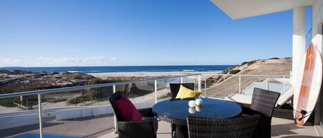 A balcony with a table and chairs, overlooking a beach and ocean.