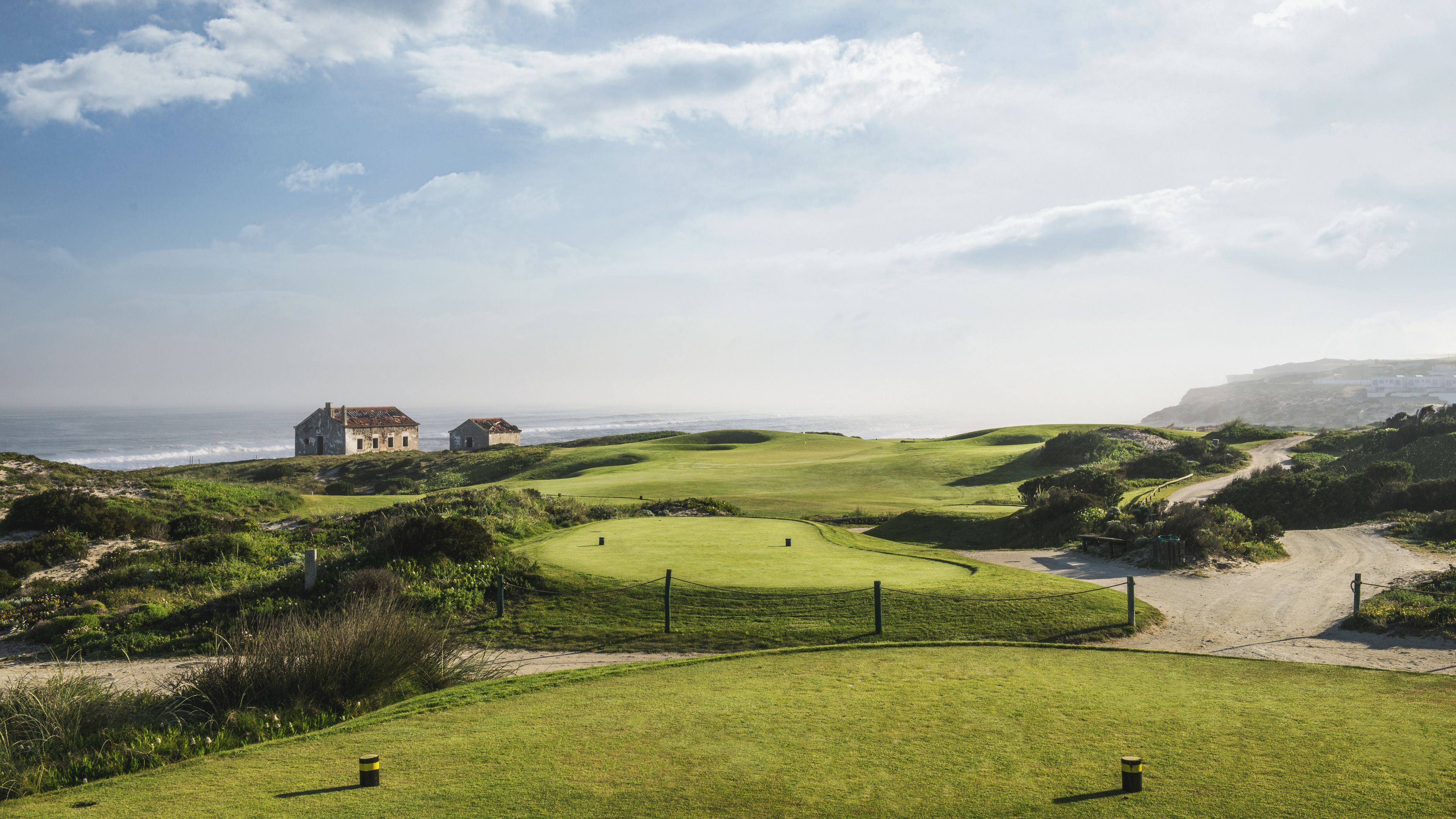 A coastal golf course with two rustic buildings, a sandy path, and a view of the ocean.