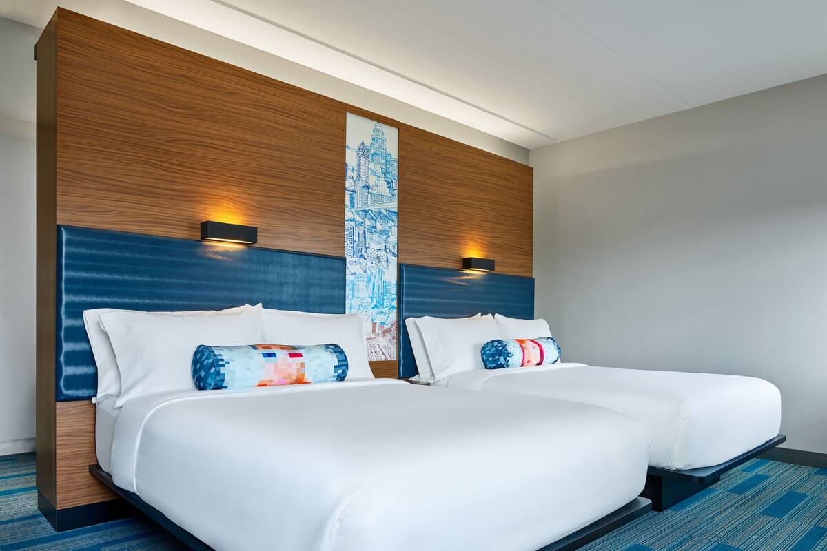 A hotel room with two beds, a wooden headboard, a cityscape artwork, and two wall-mounted lights.