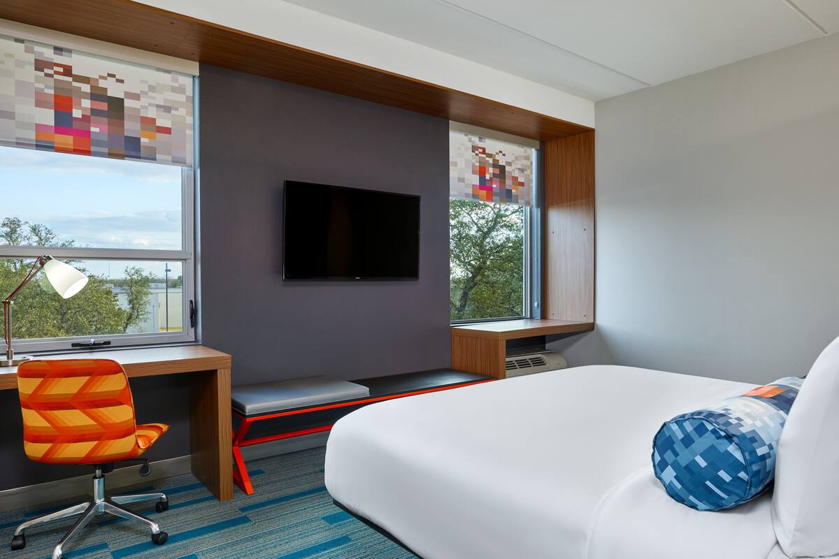 A modern hotel room with a bed, a desk, a chair, a TV, and large windows.