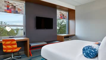 A modern hotel room with a bed, a desk, a chair, a TV, and large windows.