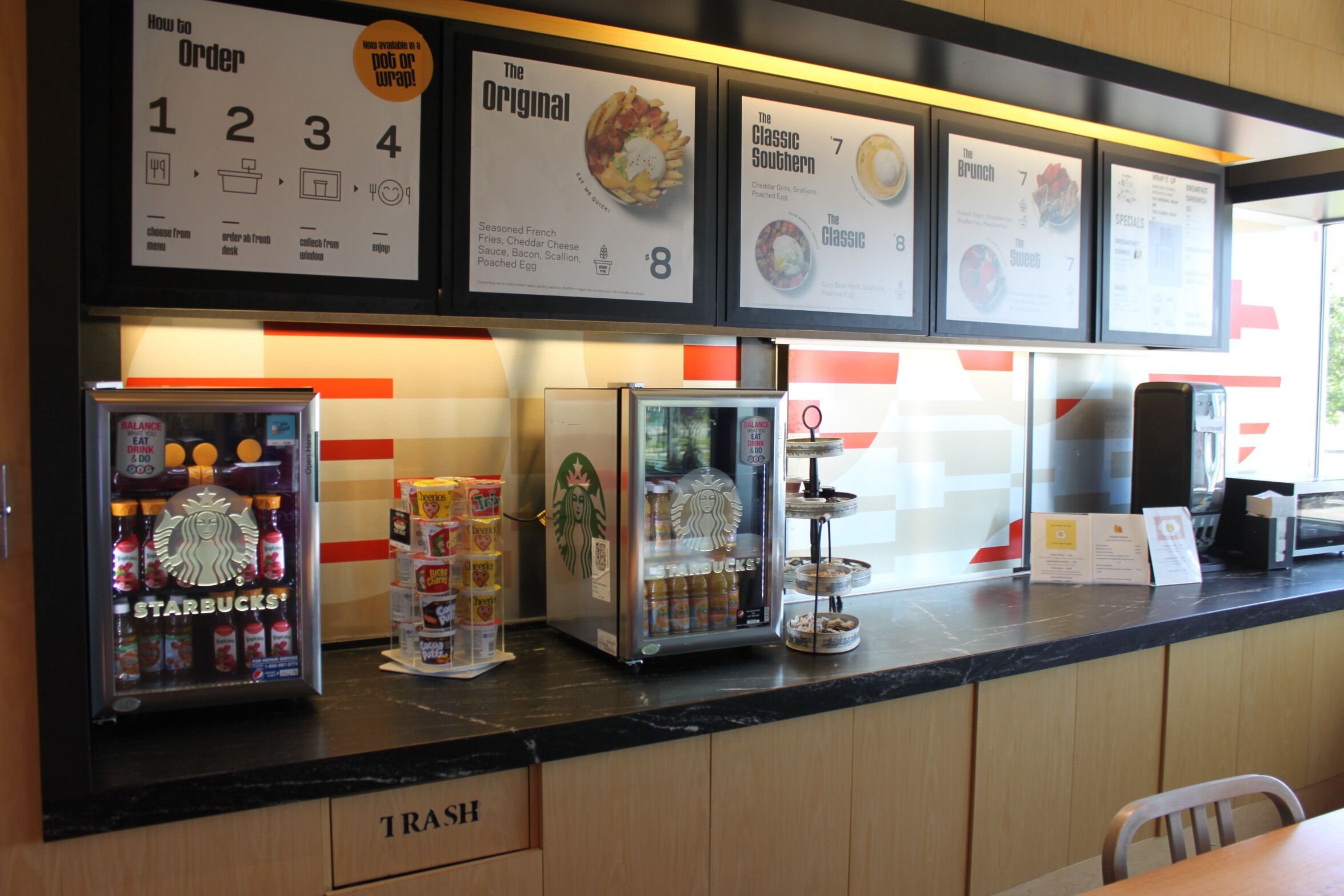 En kiosk med fastfood, menutavler, et Starbucks-køleskab og en skraldespand.