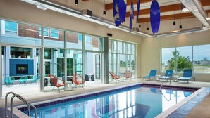 Une piscine intérieure avec des parois en verre transparent, des chaises bleues et une vue sur l’extérieur.