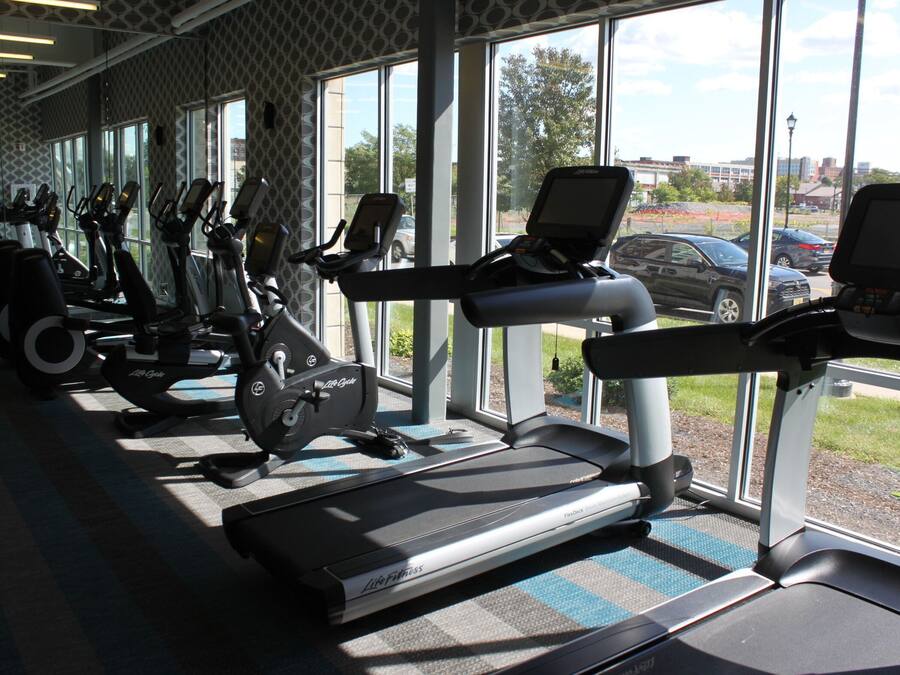 Ein Fitnessraum mit mehreren Laufbändern und Ergometer, großen Fenstern und Blick ins Freie.