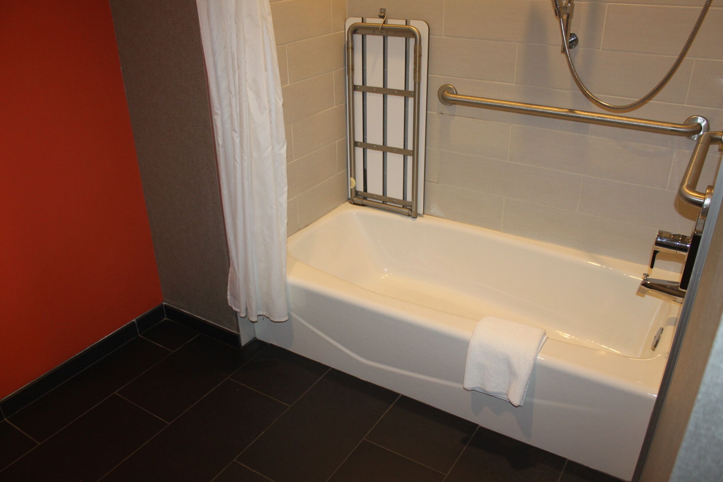 Une salle de bain d’hôtel standard, équipée d’une baignoire, de barres d’appui et d’un pommeau de douche.