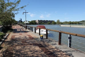 A riverside walkway with a boat dock, a red car, and a building in the background.