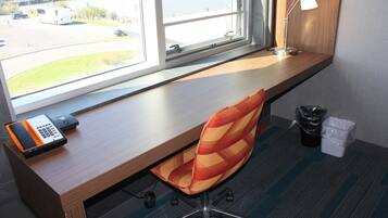 A hotel room with a desk, a chair, a lamp, a trash bin, and a window with a view of a parking lot.