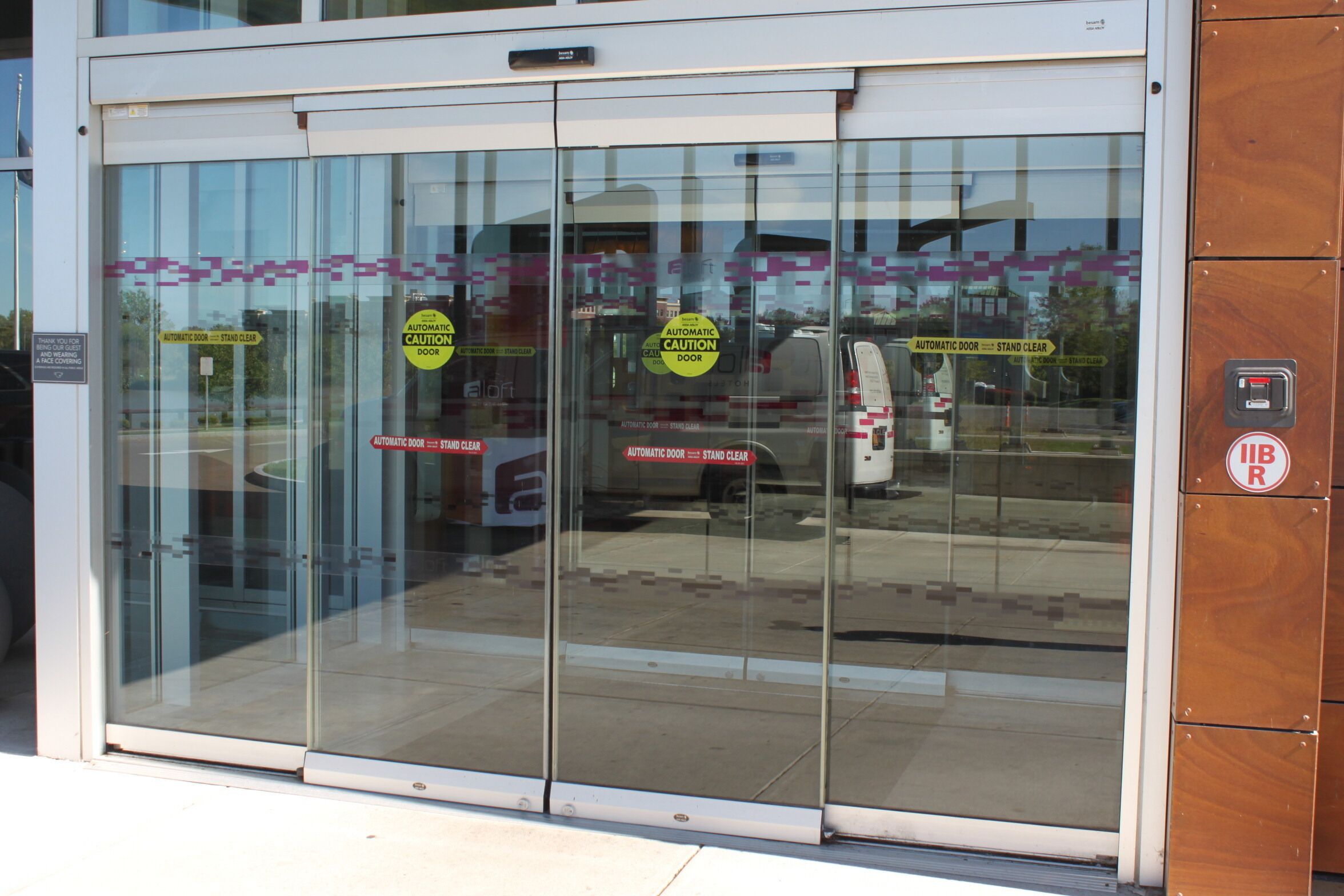 A glass door with multiple stickers indicating "Automatic Door - Stand Clear" and a "No Parking" sign.
