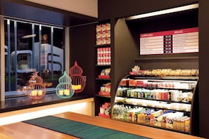 A hotel lobby with a glass partition, a menu board listing food and drink prices, and a variety of snacks and beverages on shelves.