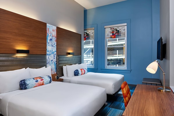 A hotel room with two beds, a wooden headboard, a large window with a view of a building, and a desk with a lamp.