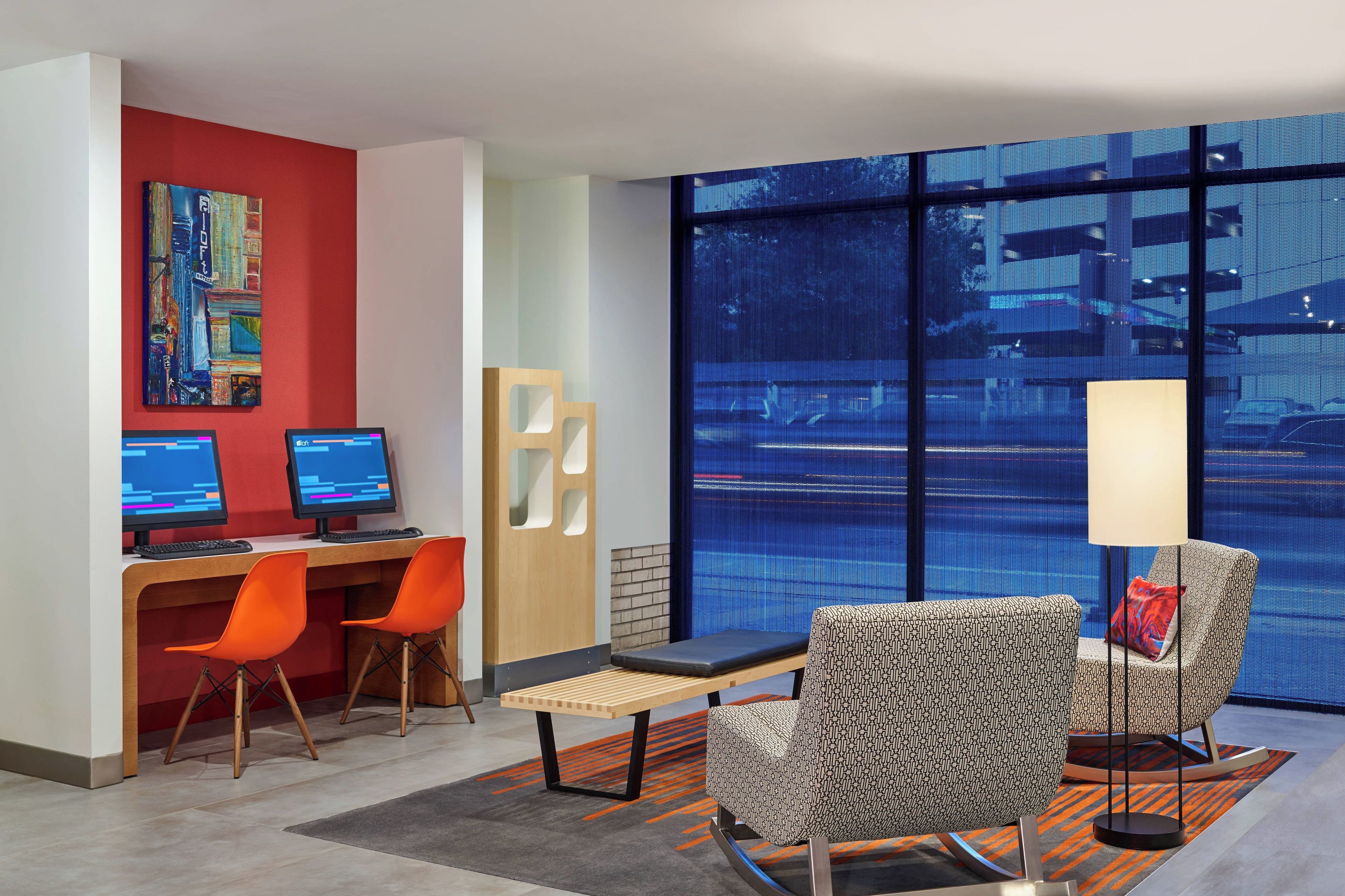 A modern hotel room with two workstations, a seating area with patterned chairs, a large window with a view, and a painting on the wall.