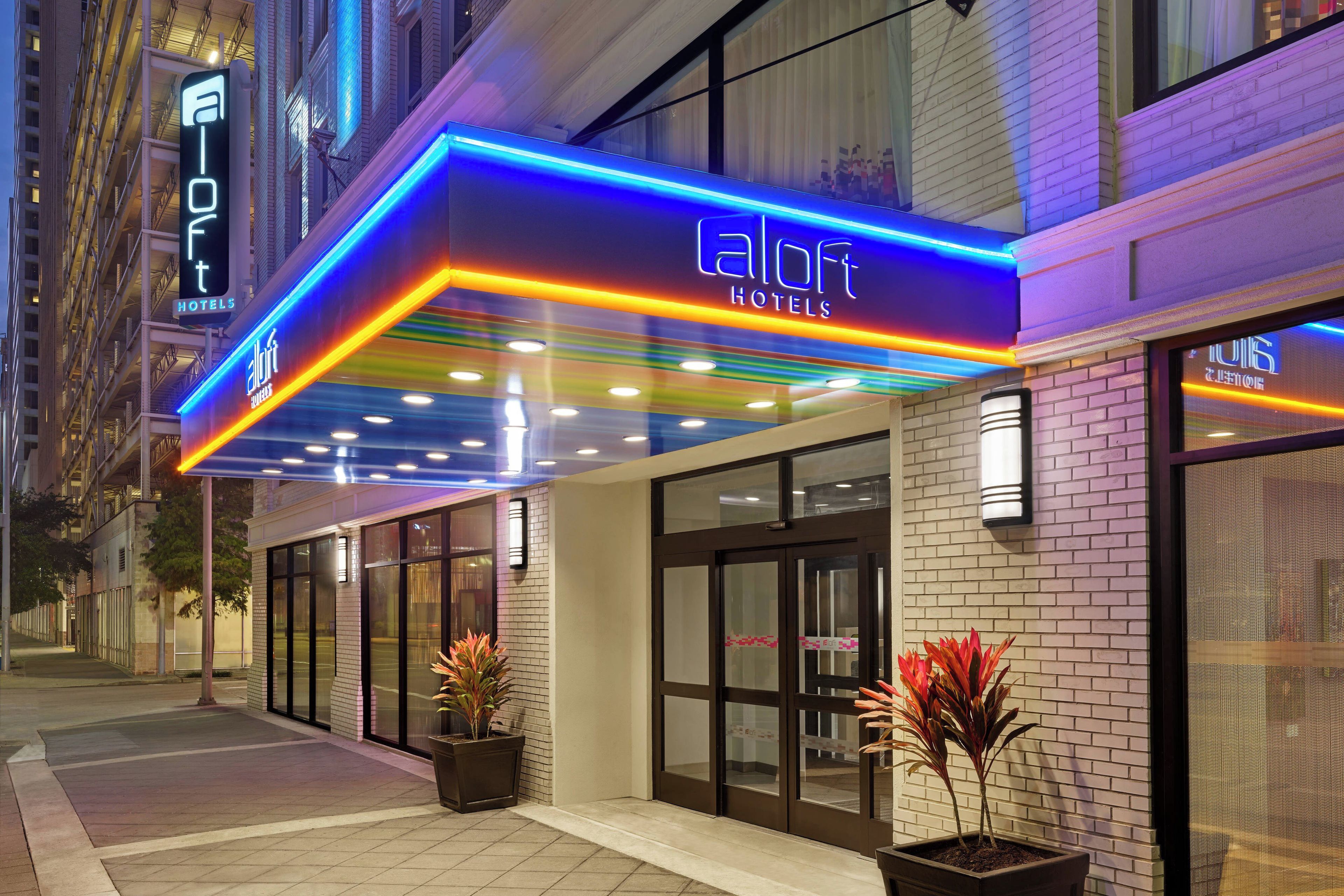 A hotel entrance with a neon sign, glass doors, and potted plants.