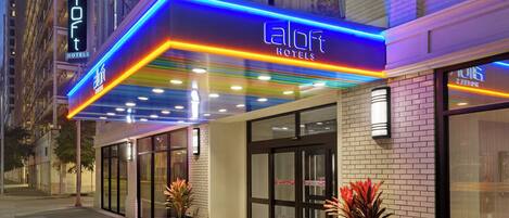A hotel entrance with a neon sign, glass doors, and potted plants.