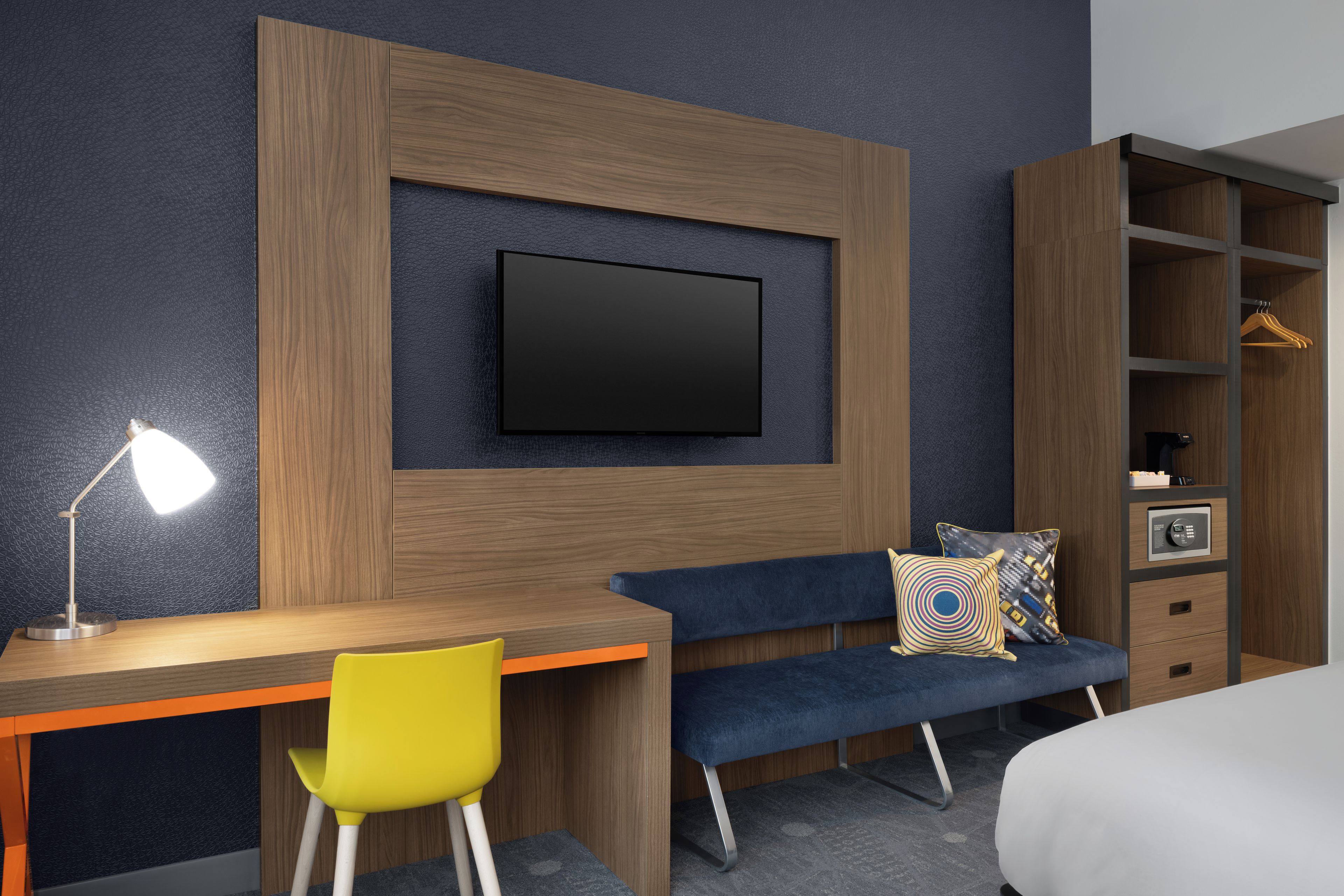 Een moderne hotelkamer met een houten bureau, een blauwe bank, een aan de muur bevestigde televisie en een kledingkast met hangruimte.