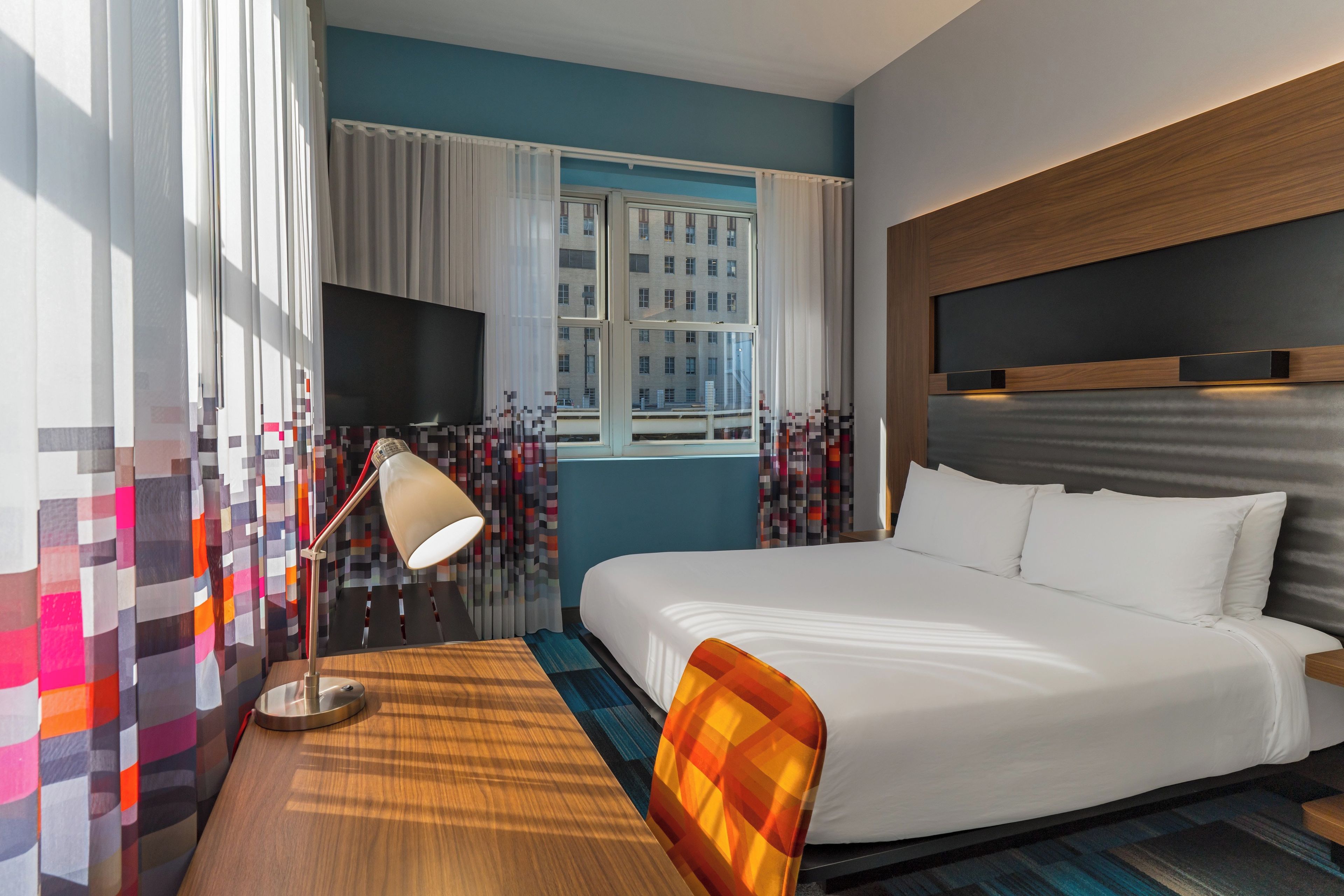 Une chambre d’hôtel avec un lit, un bureau, une chaise, une télévision et une fenêtre avec des rideaux.