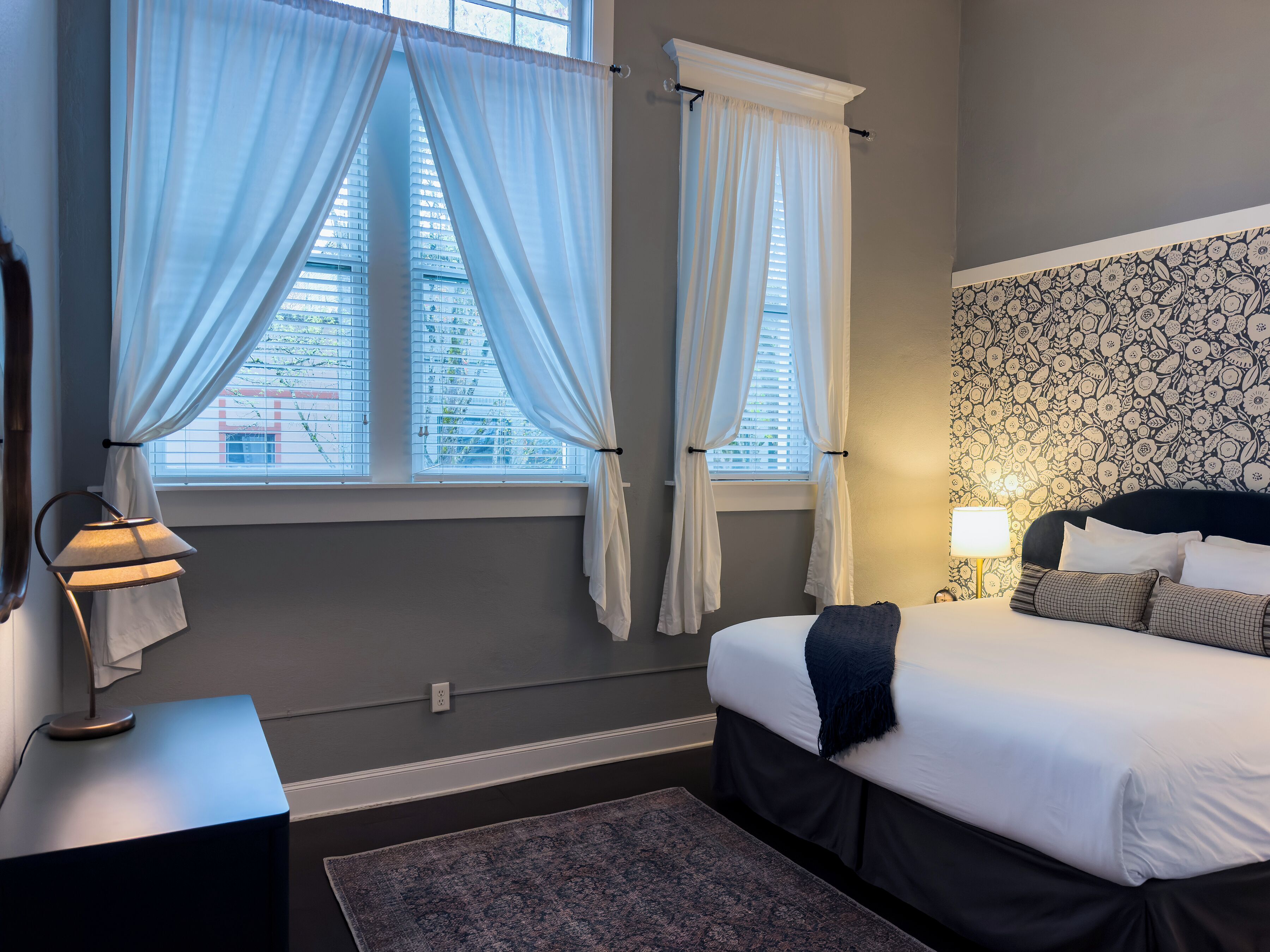A bedroom with a bed, two windows with curtains, a bedside lamp, a patterned wall, and a rug.