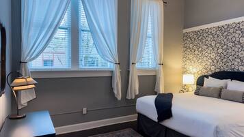 A bedroom with a bed, two windows with curtains, a bedside lamp, a patterned wall, and a rug.