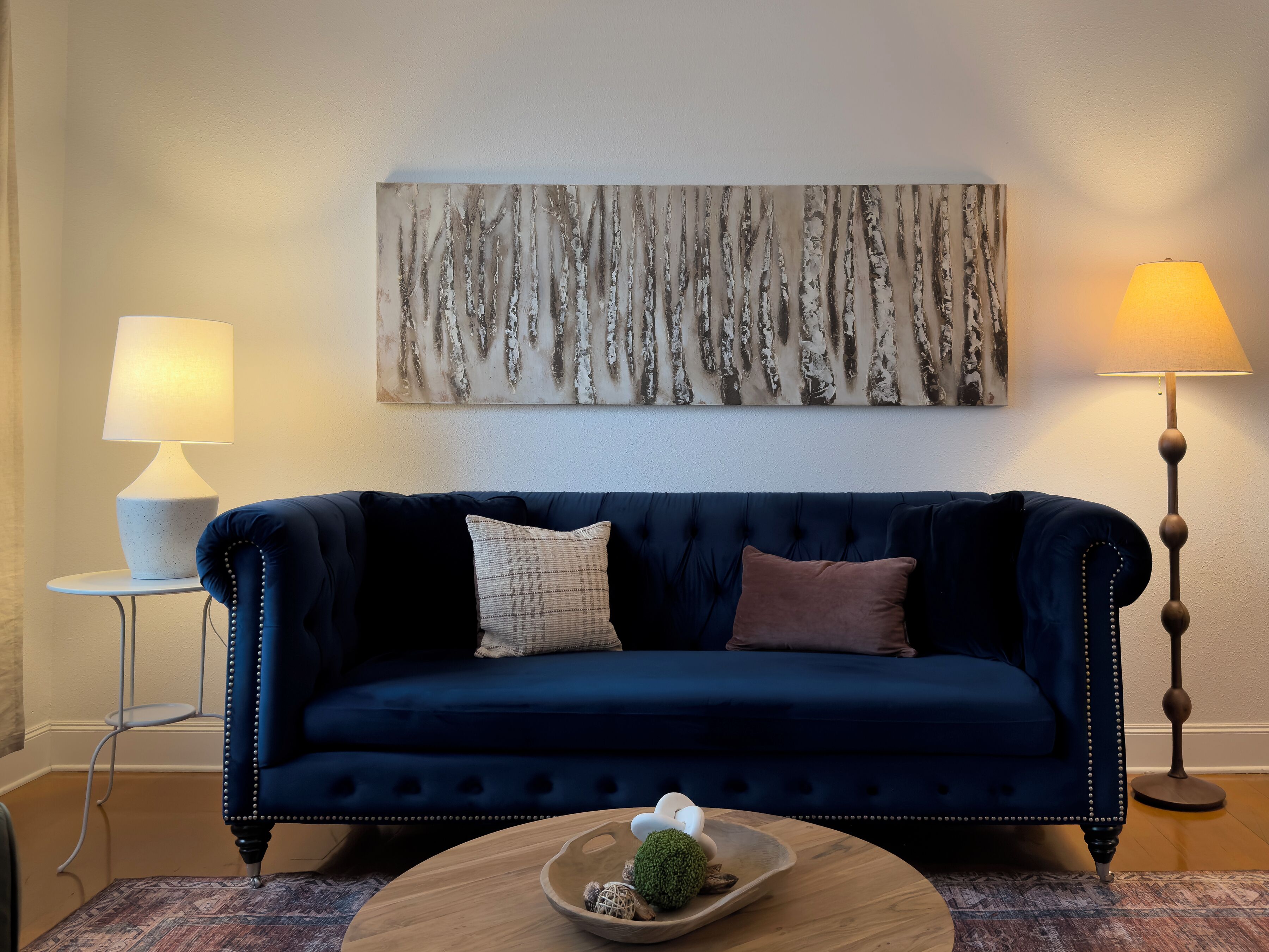 A dark blue upholstered sofa with tufted detailing, a round wooden coffee table with decorative items, a wall-mounted abstract painting, and two floor lamps.