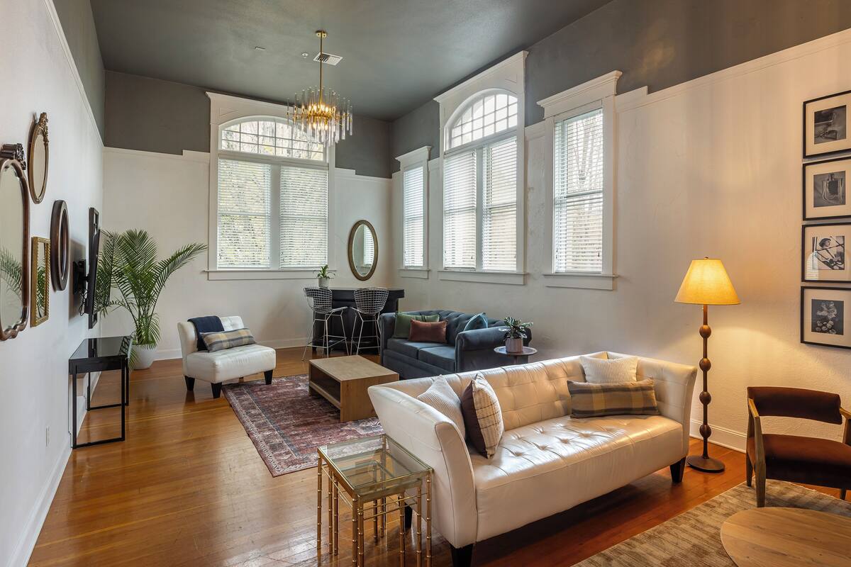 A living room with a sofa, coffee table, and lamp; a dining area with chairs and a table; and a large window with blinds.