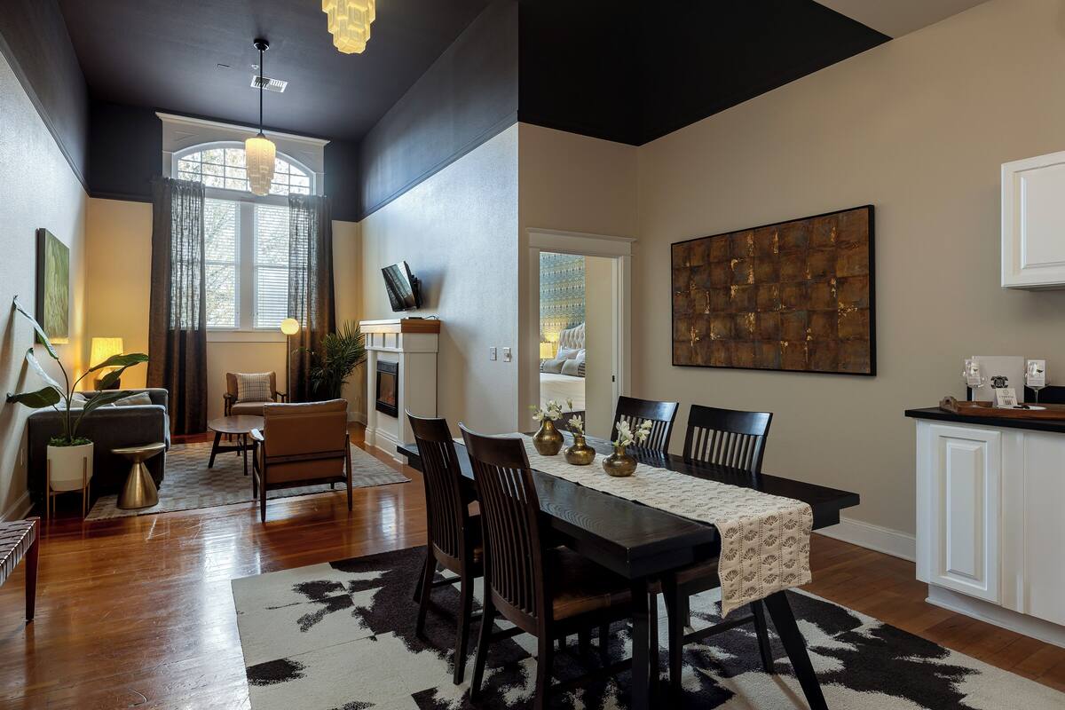 A dining area with a table set for four, surrounded by chairs, a patterned rug, and a large abstract painting on the wall.