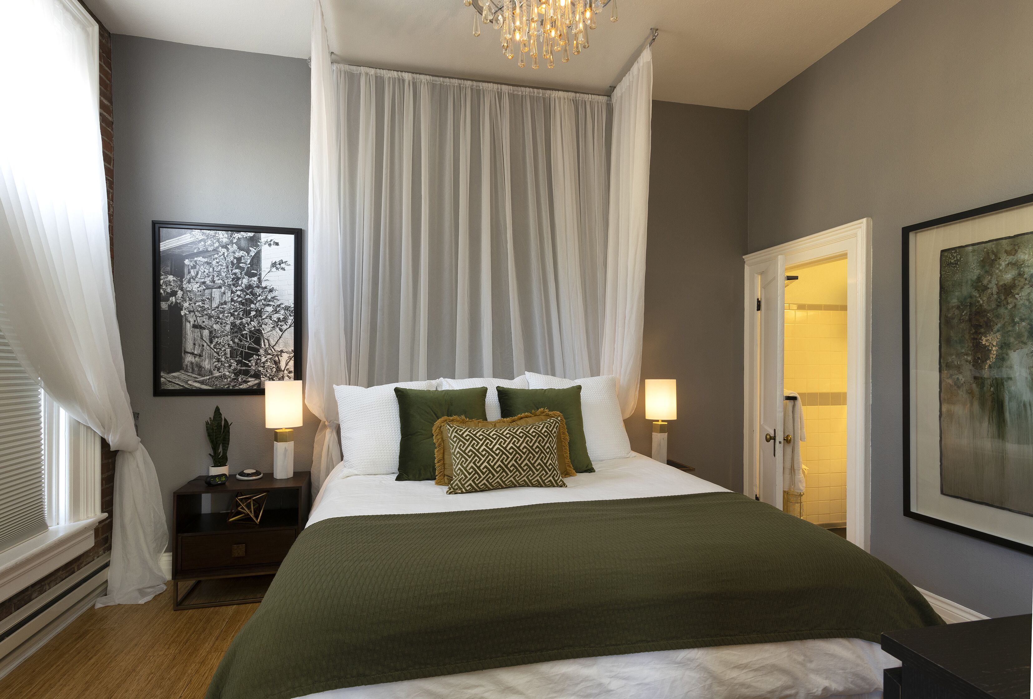 A bedroom with a canopy bed, bedside lamps, a framed picture on the wall, and a door leading to a bathroom.