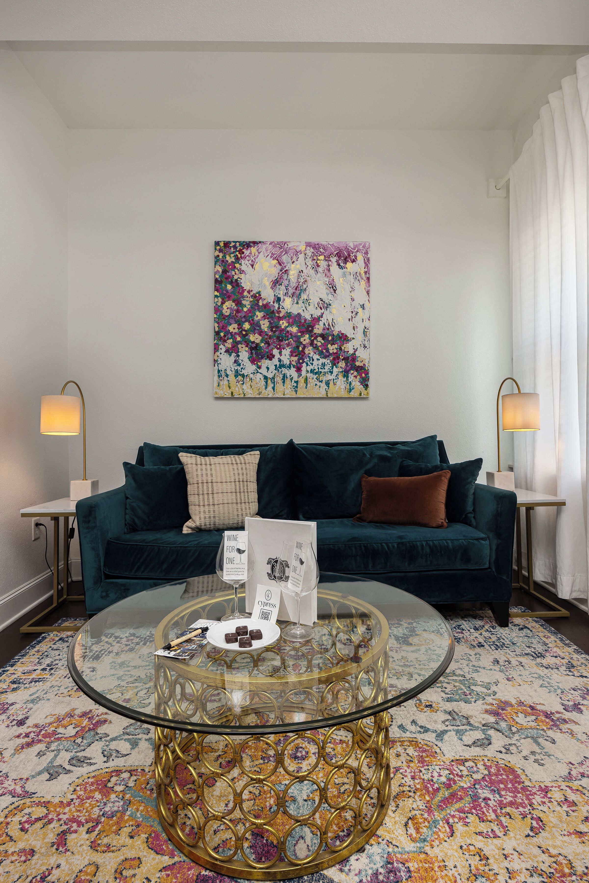 Ein Wohnzimmer mit einem dunkelgrünen Sofa, einem Glastisch mit goldfarbener Basis, einem floralen Teppich und einem farbenfrohen, abstrakten Gemälde an der Wand.