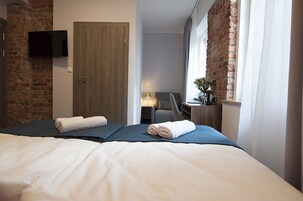 A hotel room with a brick wall, a wooden door, a bed with white linens, a flat-screen TV mounted on the wall, and a window with curtains.