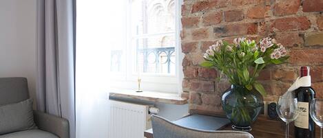A hotel room with a brick wall, a window with curtains, a grey sofa, a wooden desk, a vase with flowers, and a wine bottle.