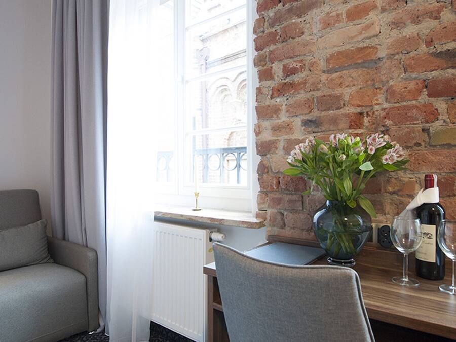 Hotellihuone, jossa on tiiliseinä, ikkuna, jossa on verhot, harmaa sohva, puinen työpöytä, maljakko kukkineen ja viinipullo.