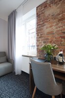 Una camera d'albergo con una parete in mattoni, una finestra con tende, un divano grigio, una scrivania in legno, un vaso con fiori e una bottiglia di vino.