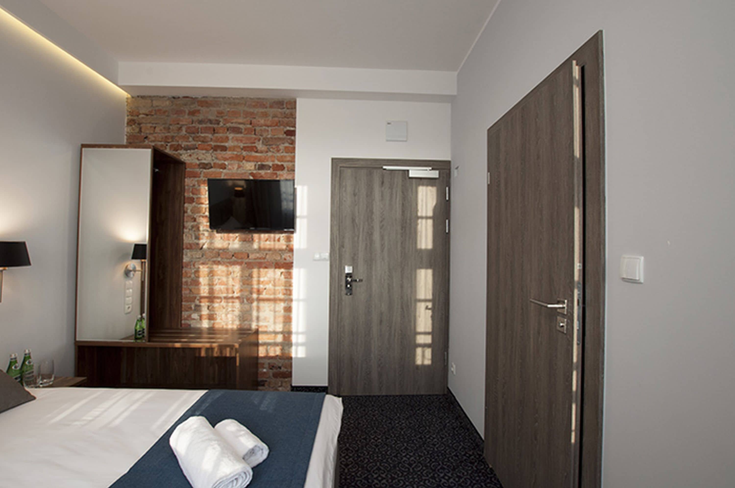 A hotel room with a brick wall, a flat-screen TV, a wooden door, and a bed with a blue bedspread and white linens.