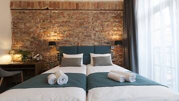 A double bed with two pillows and rolled towels, a brick wall, a nightstand with a lamp, and a window with curtains.